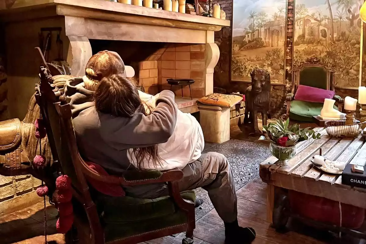 Dos personas sentadas abrazadas en una silla frente a una chimenea en una sala acogedora decorada con velas, cojines y una mesa de madera Dos personas sentadas abrazadas en una silla frente a una chimenea en una sala acogedora decorada con velas, cojines y una mesa de madera