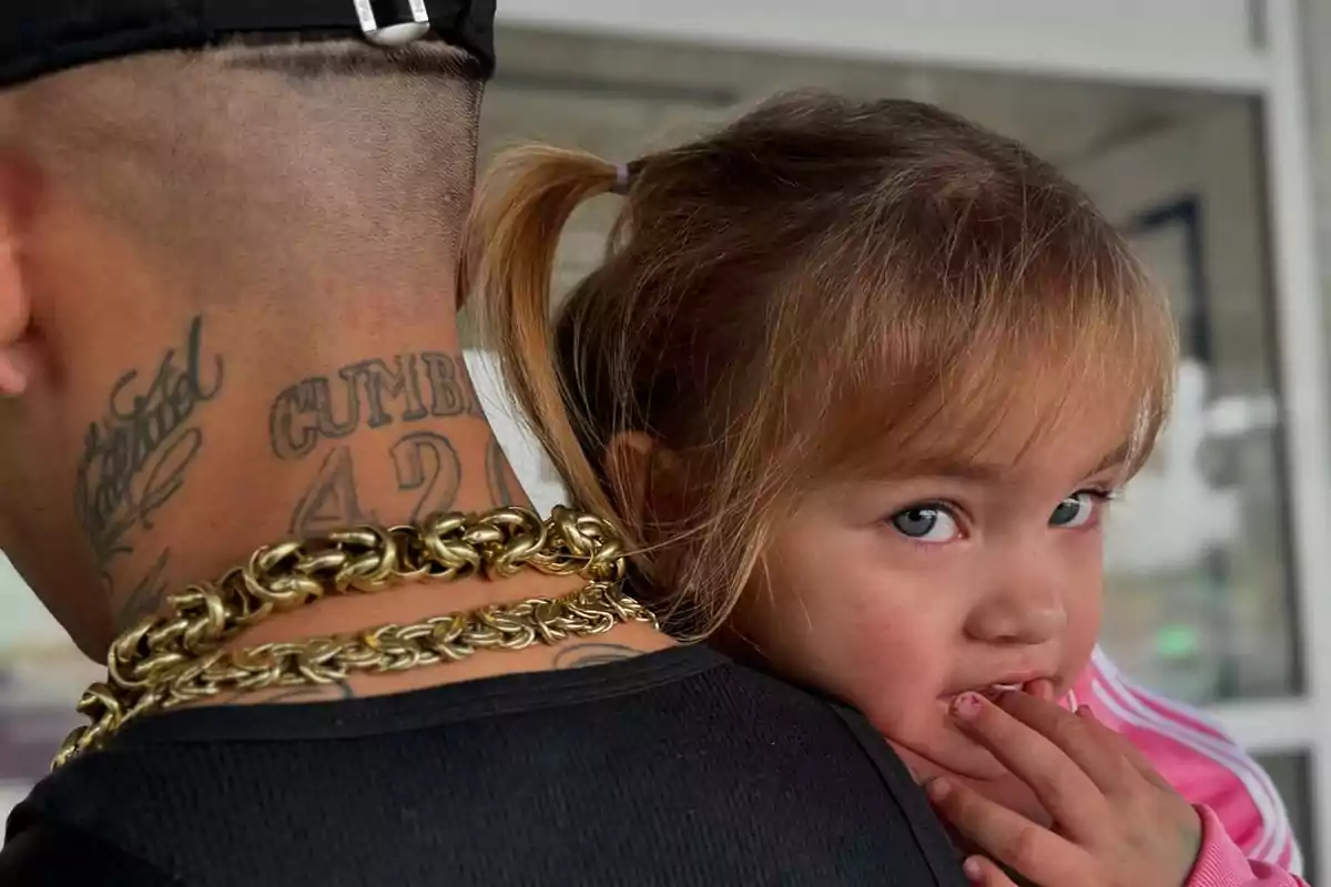 A little girl with light eyes and blonde hair tied back leans on the shoulder of a man with tattoos on his neck and gold chains. A little girl with light eyes and blonde hair tied back leans on the shoulder of a man with tattoos on his neck and gold chains.