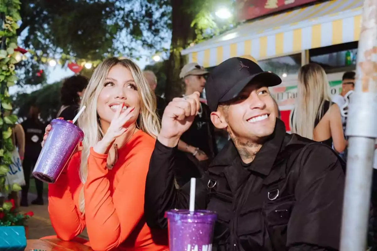 Dos personas sonrientes sentadas en una mesa al aire libre, una mujer con un vestido naranja sostiene un vaso brillante y un hombre con gorra negra también sostiene un vaso similar, con un fondo de luces y vegetación.