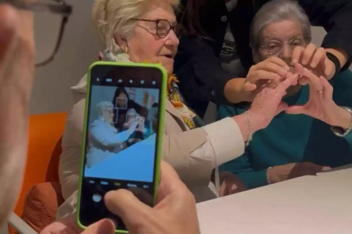 Personas mayores sentadas en una mesa formando un corazón con las manos mientras alguien toma una foto con un teléfono móvil Personas mayores sentadas en una mesa formando un corazón con las manos mientras alguien toma una foto con un teléfono móvil