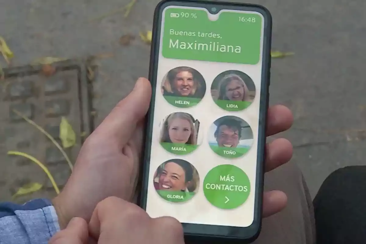 Persona sosteniendo un teléfono móvil que muestra una aplicación con fotos de contactos y el saludo Buenas tardes Maximiliana en la pantalla Persona sosteniendo un teléfono móvil que muestra una aplicación con fotos de contactos y el saludo Buenas tardes Maximiliana en la pantalla