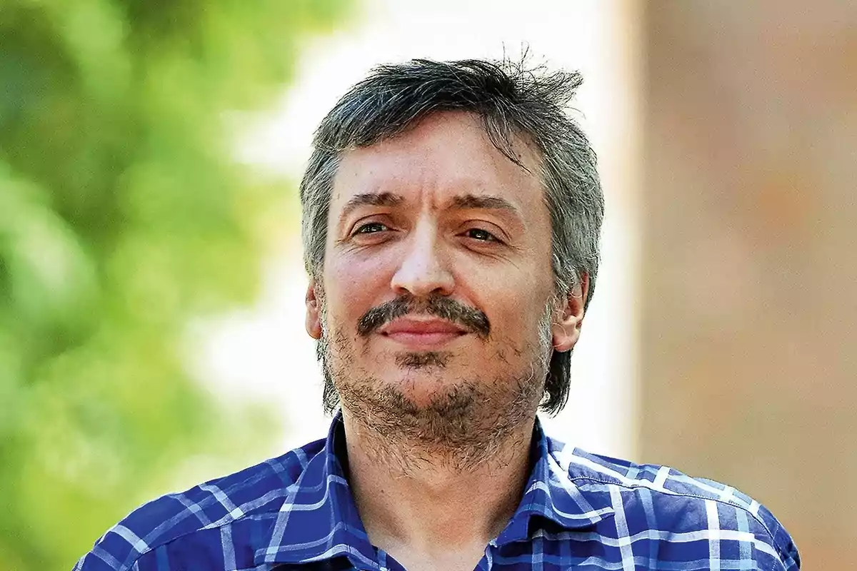 Un hombre con camisa a cuadros azules y barba ligera, con un fondo desenfocado de vegetación.