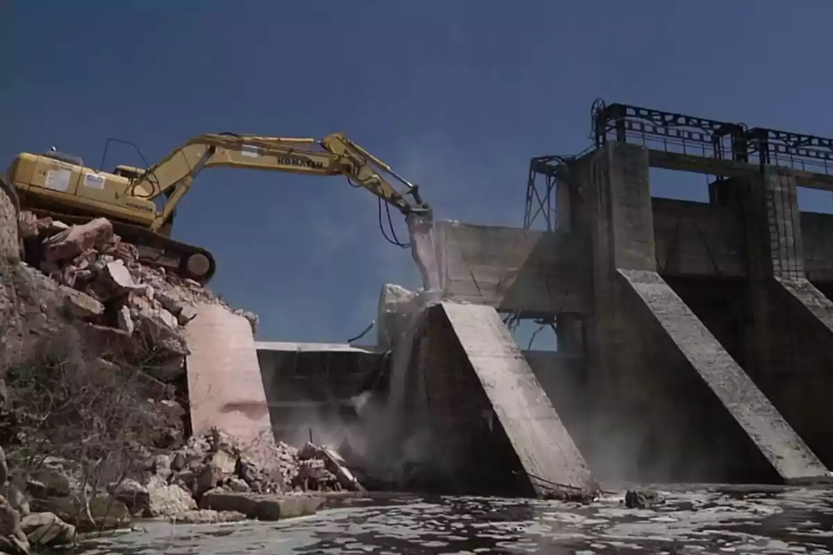 Una excavadora demoliendo una estructura de concreto junto a un cuerpo de agua bajo un cielo despejado.