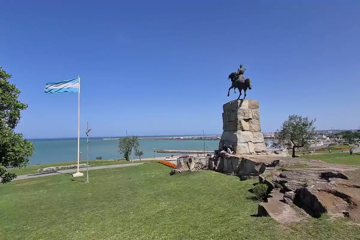 An equestrian statue on a stone pedestal with a flag waving nearby and the sea in the background on a sunny day.