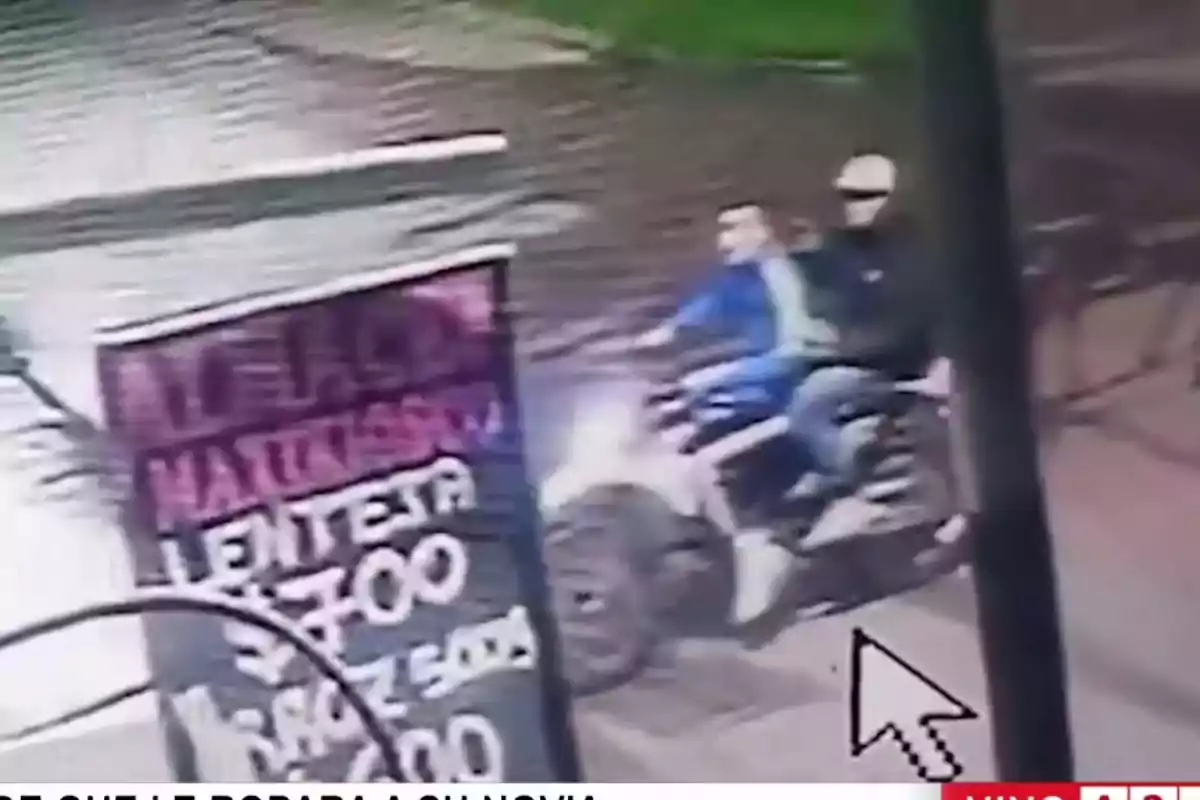Dos personas en una motocicleta pasan junto a un cartel en una calle. Dos personas en una motocicleta pasan junto a un cartel en una calle.