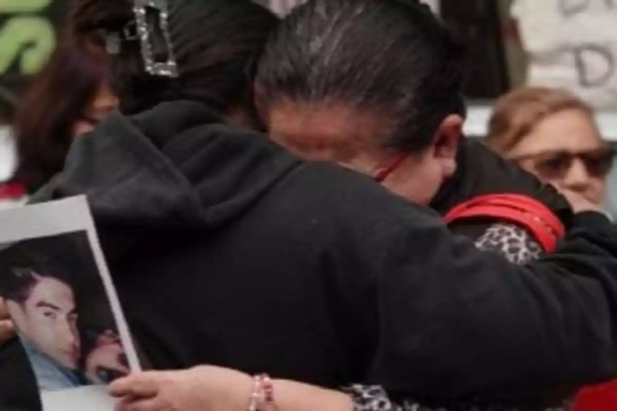 Dos personas se abrazan mientras una sostiene una fotografía en un entorno que sugiere una reunión emotiva. Dos personas se abrazan mientras una sostiene una fotografía en un entorno que sugiere una reunión emotiva.