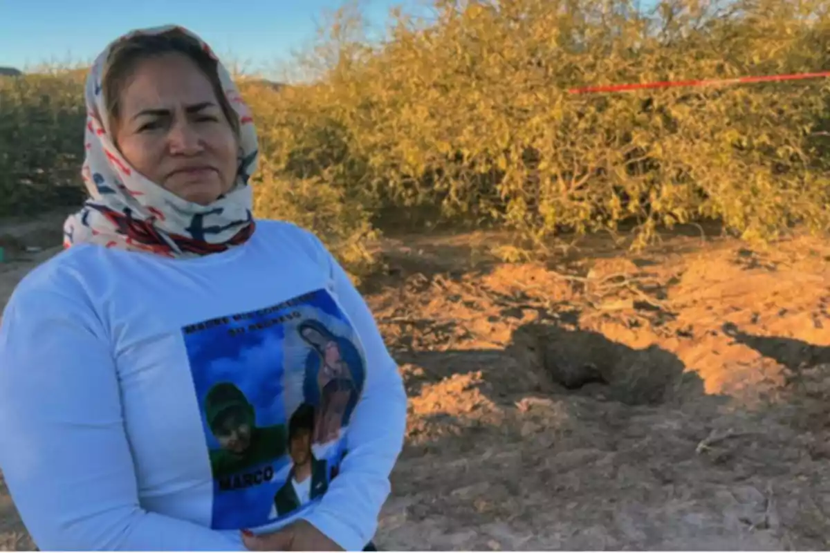 Una mujer con un pañuelo en la cabeza y una camiseta con imágenes impresas está de pie en un área desértica con arbustos y un agujero en el suelo. Una mujer con un pañuelo en la cabeza y una camiseta con imágenes impresas está de pie en un área desértica con arbustos y un agujero en el suelo.