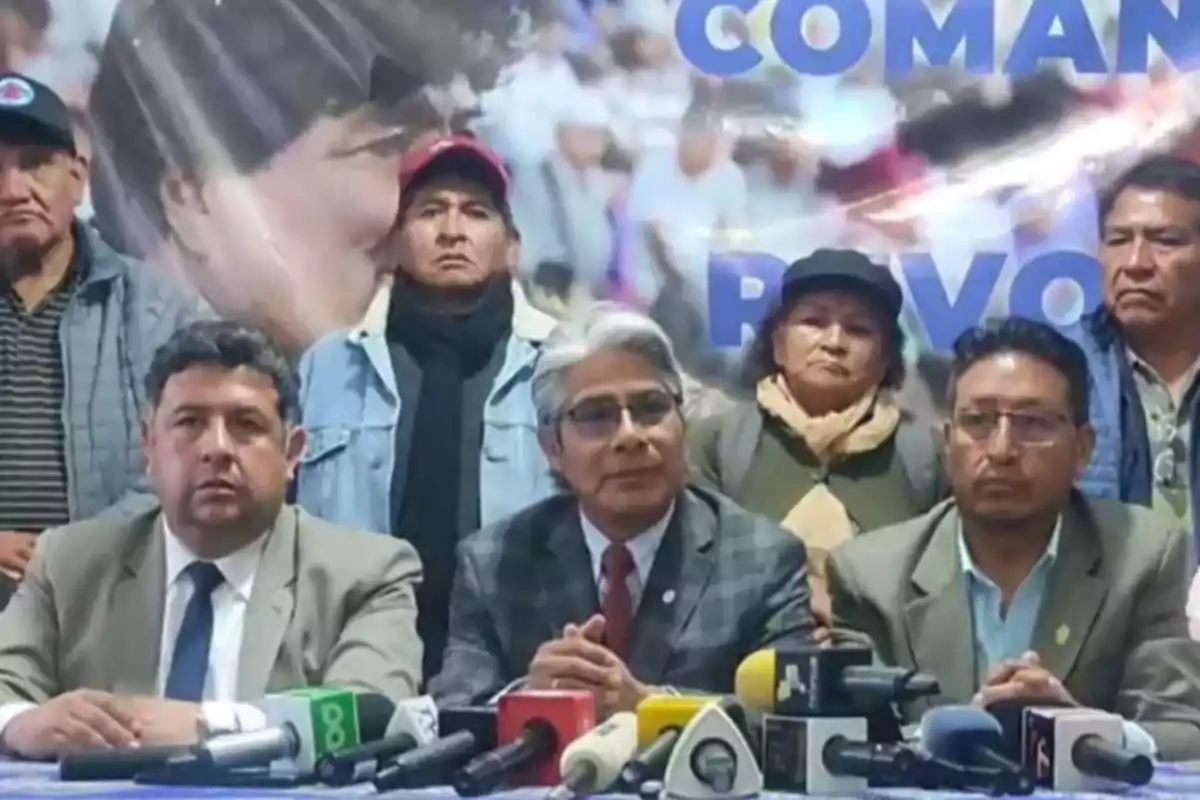 Un grupo de personas está sentado frente a varios micrófonos en una conferencia de prensa con un cartel de fondo. Un grupo de personas está sentado frente a varios micrófonos en una conferencia de prensa con un cartel de fondo.