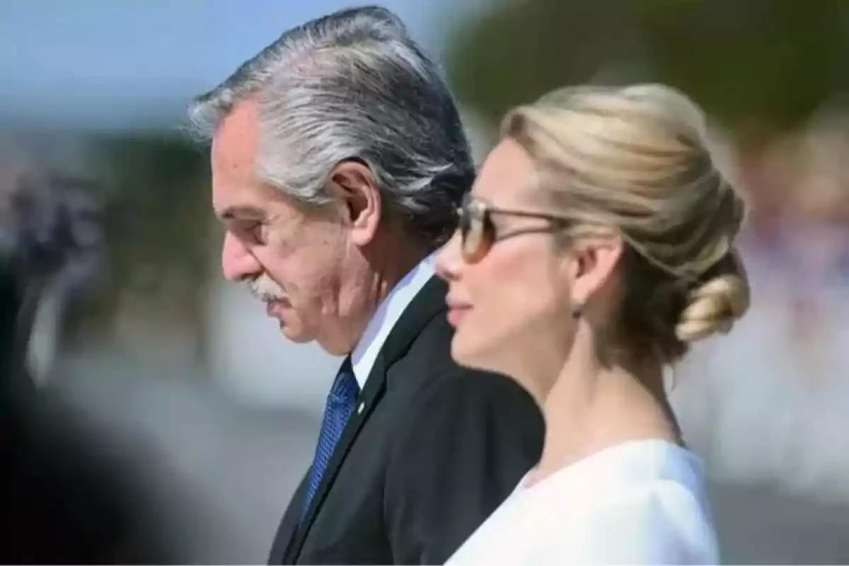 Dos personas de perfil en un evento formal al aire libre, una de ellas lleva traje y la otra viste de blanco y usa gafas de sol Dos personas de perfil en un evento formal al aire libre, una de ellas lleva traje y la otra viste de blanco y usa gafas de sol