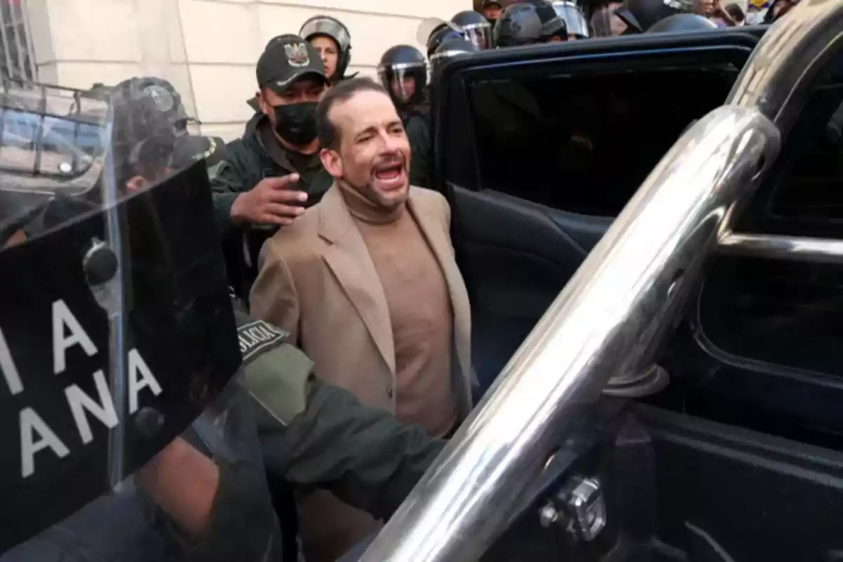 A man is being escorted by police officers to a vehicle while a crowd watches.