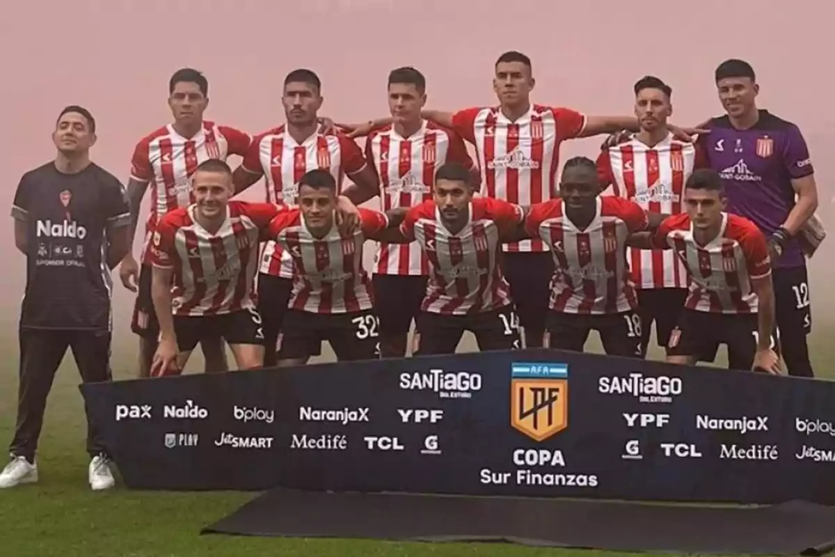 Un equipo de fútbol posando para una foto grupal en el campo, con camisetas a rayas rojas y blancas, detrás de un cartel con logotipos de patrocinadores.