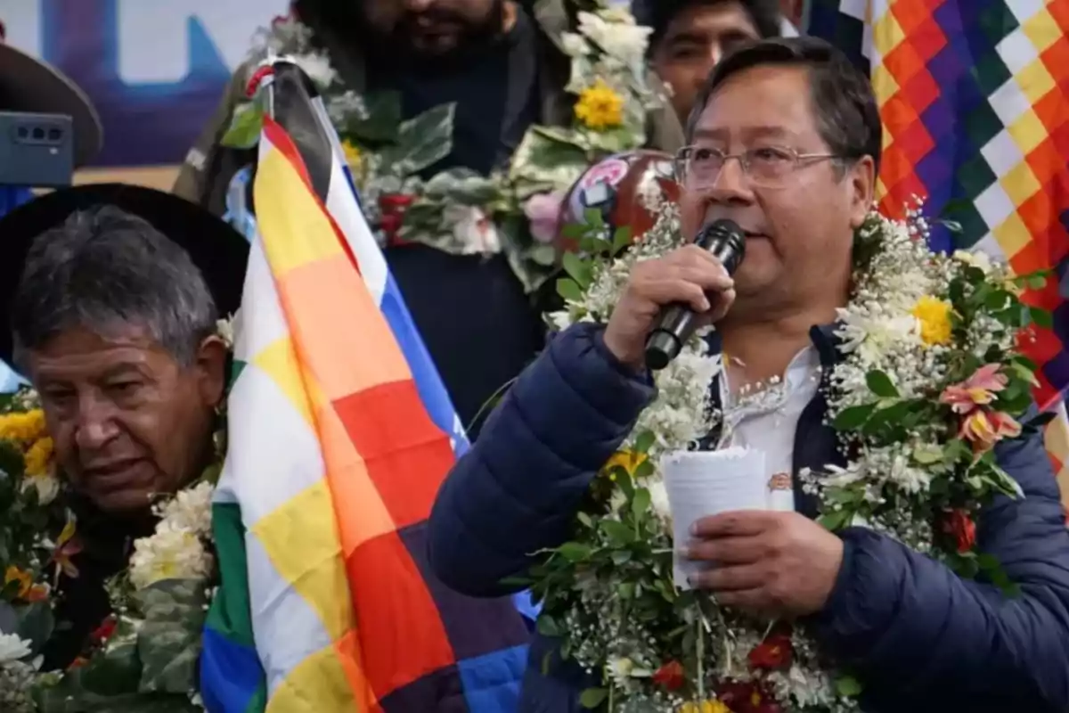 Un hombre con gafas y una guirnalda de flores habla por un micrófono mientras sostiene una hoja de papel, junto a él hay una bandera colorida y otra persona con una guirnalda similar.