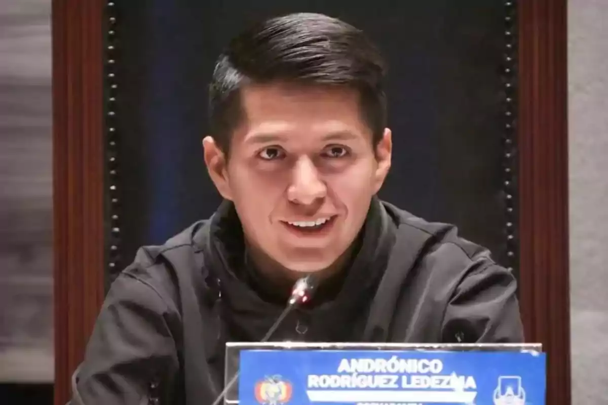 A young man with short dark hair smiles while sitting in front of a microphone and a nameplate on a table. A young man with short dark hair smiles while sitting in front of a microphone and a nameplate on a table.