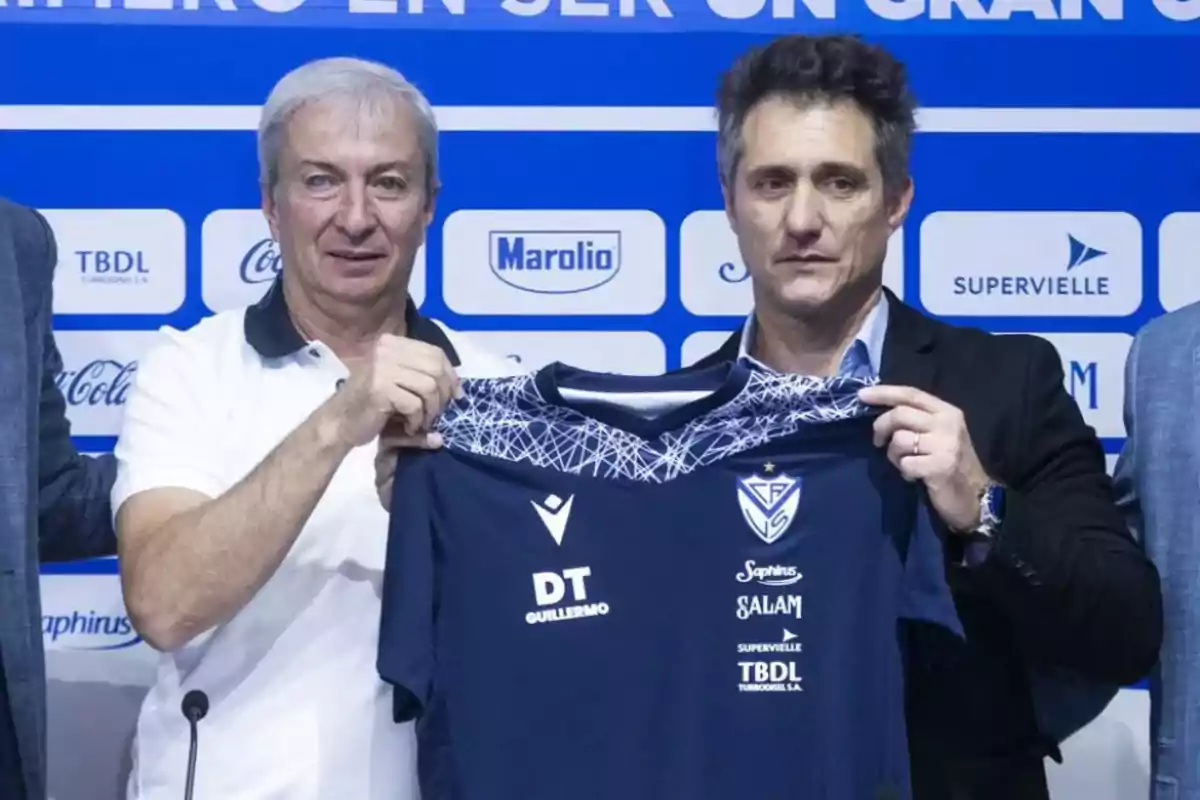 Two men pose together holding a blue soccer team jersey in front of a backdrop with sponsor logos. Two men pose together holding a blue soccer team jersey in front of a backdrop with sponsor logos.