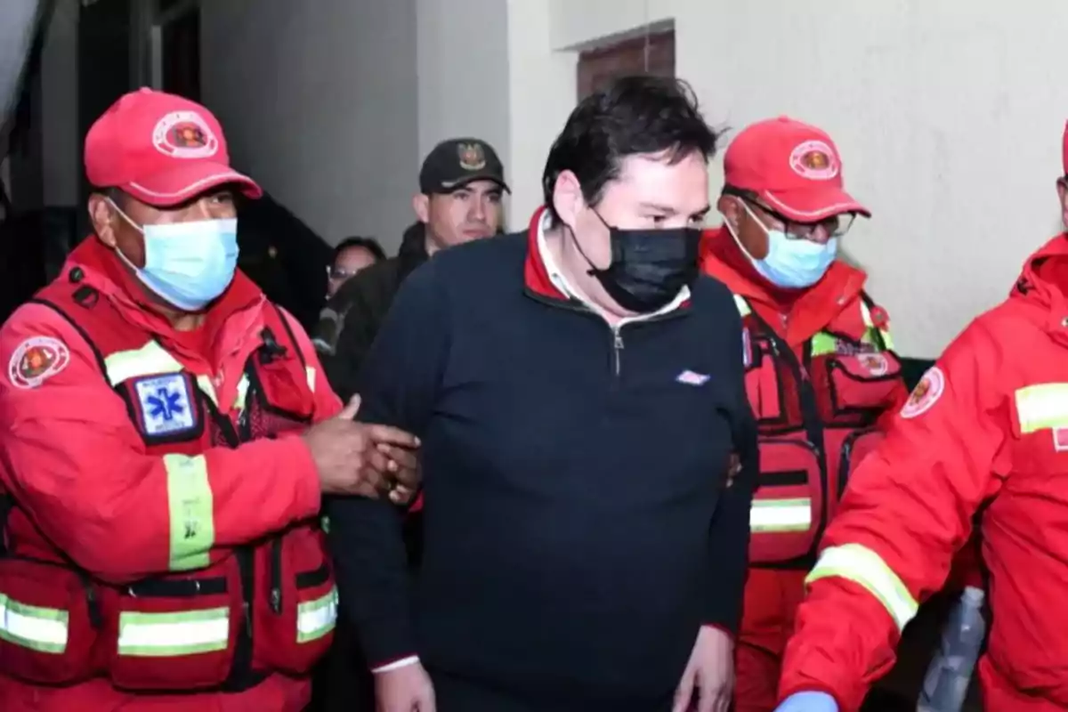 Un hombre con mascarilla negra es escoltado por varios rescatistas vestidos con uniformes rojos y chalecos reflectantes Un hombre con mascarilla negra es escoltado por varios rescatistas vestidos con uniformes rojos y chalecos reflectantes