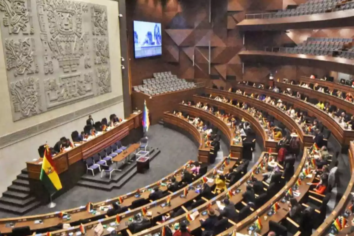 Una sala de asambleas con un diseño semicircular, donde se encuentran varias personas sentadas en escritorios, con una gran bandera y un mural decorativo en la pared.