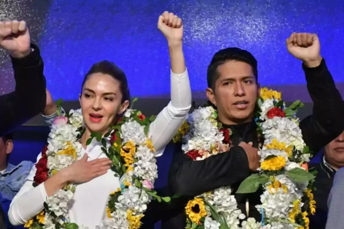 Two people with flower garlands raise their fists and place their other hand on their chest at a public event. Two people with flower garlands raise their fists and place their other hand on their chest at a public event.