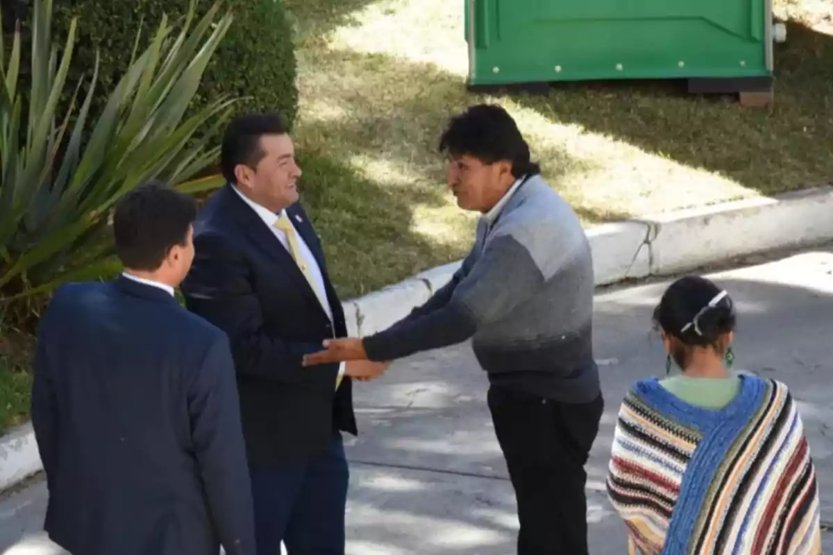 Four people are talking outdoors; two of them are shaking hands while another person watches, and a woman wearing a colorful shawl has her back turned. Four people are talking outdoors; two of them are shaking hands while another person watches, and a woman wearing a colorful shawl has her back turned.