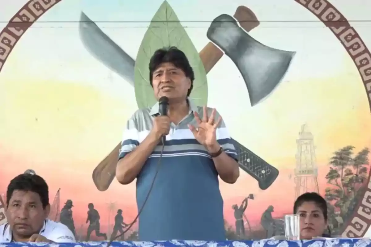 A man speaking into a microphone on a stage with a mural in the background showing a leaf and farming tools, accompanied by two people sitting beside him. A man speaking into a microphone on a stage with a mural in the background showing a leaf and farming tools, accompanied by two people sitting beside him.