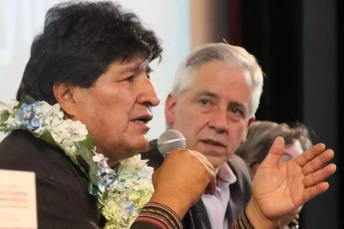 Man with a flower garland speaking into a microphone while another person watches him closely at a public event