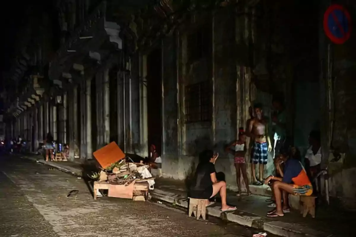 Una calle oscura con personas sentadas y de pie junto a un edificio deteriorado, con escombros en la acera.