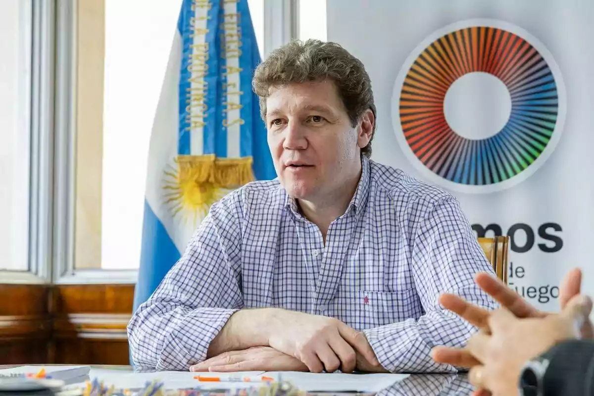 A man in a checkered jersey is sitting at a table with papers, behind him there is an Argentine flag and a sign with a colorful circular design.