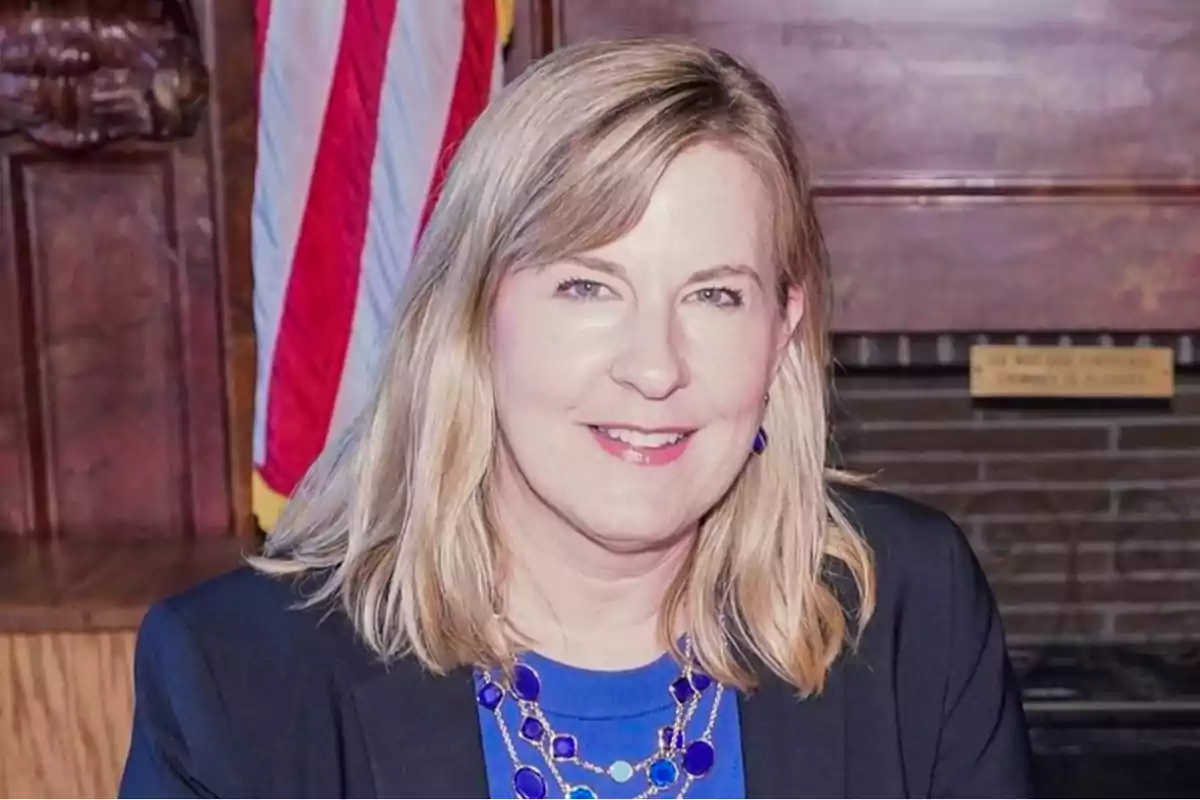 Blonde woman with a blue necklace and dark jacket smiling in front of a United States flag