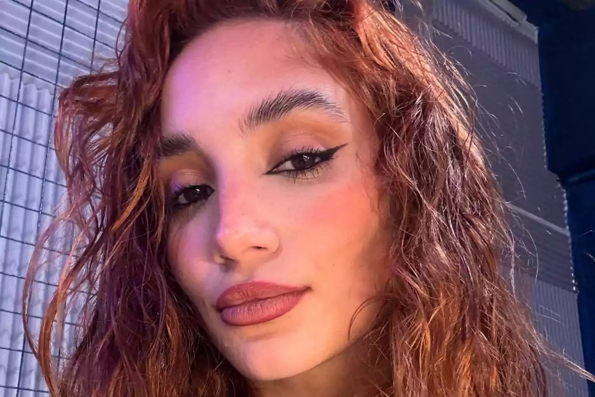A person with curly hair and eye makeup, posing in front of a metal fence.