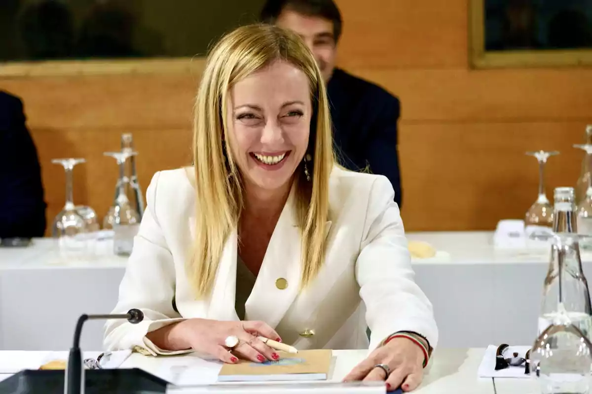 Una mujer sonriente con cabello rubio y chaqueta blanca está sentada en una mesa de conferencia con botellas y copas de vidrio.