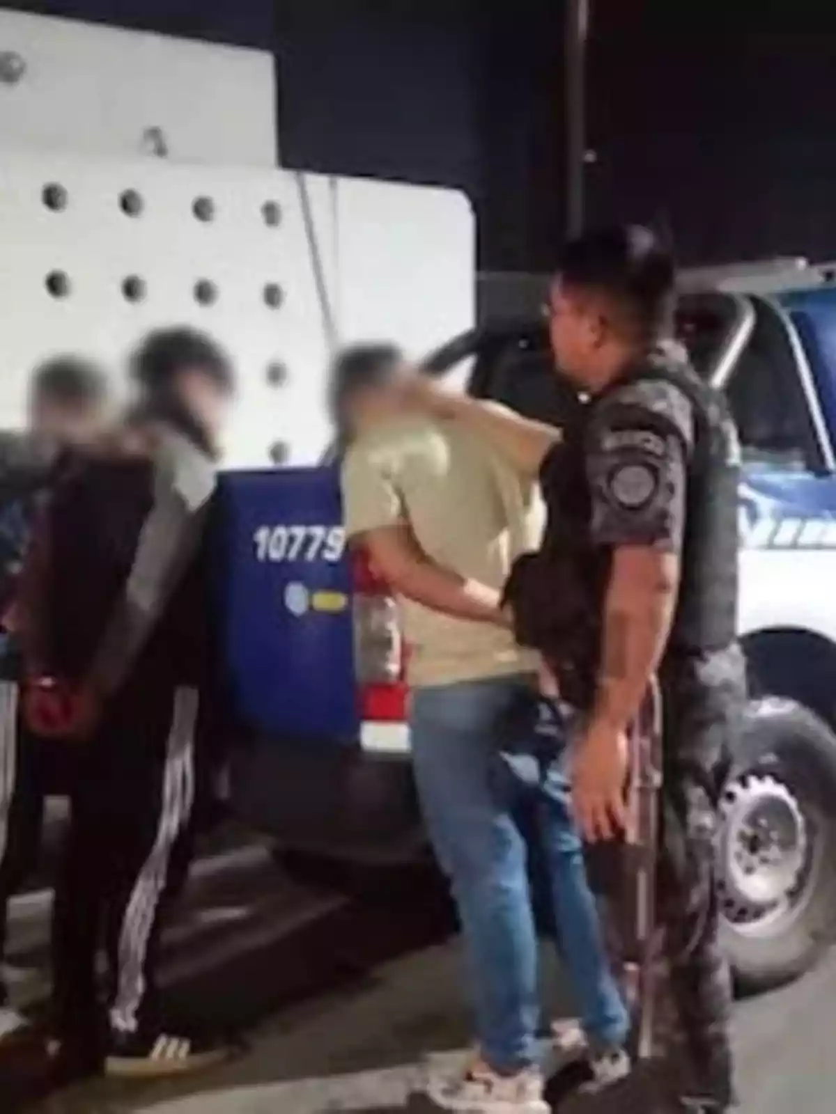 A group of people handcuffed next to a patrol car while a police officer watches the situation.