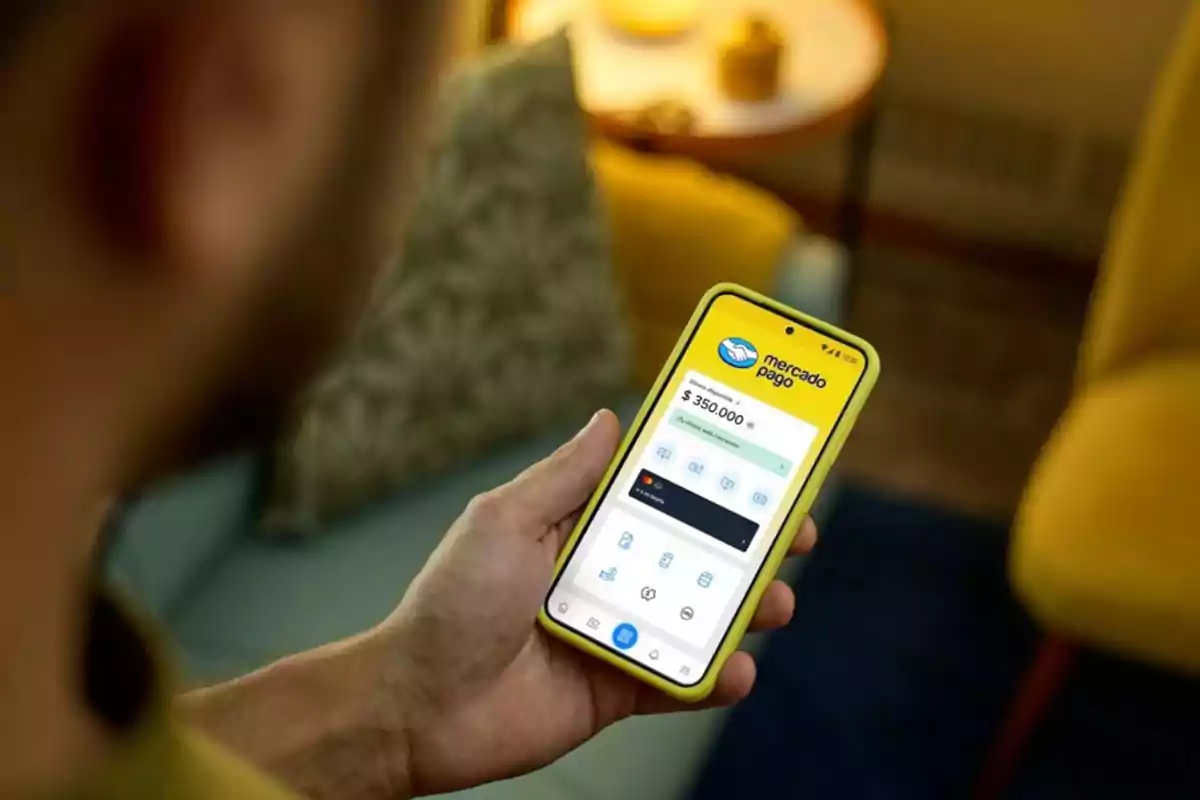 Persona sosteniendo un teléfono móvil con la aplicación de Mercado Pago abierta en la pantalla en un ambiente hogareño con sillones y una mesa de fondo Persona sosteniendo un teléfono móvil con la aplicación de Mercado Pago abierta en la pantalla en un ambiente hogareño con sillones y una mesa de fondo