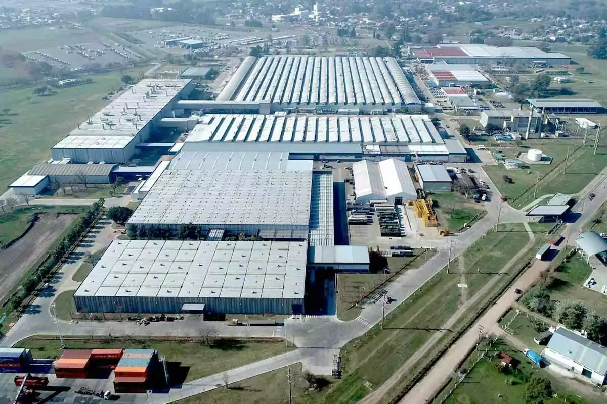 Aerial view of a large industrial complex with several buildings and metal roofs surrounded by green areas and paved roads Aerial view of a large industrial complex with several buildings and metal roofs surrounded by green areas and paved roads
