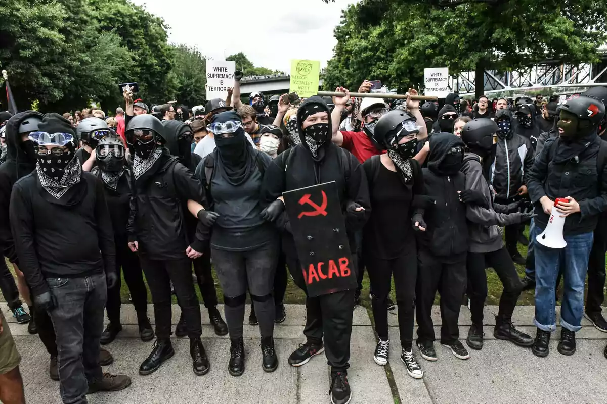 Un grupo de personas vestidas de negro con máscaras y cascos se manifiestan al aire libre, algunas sostienen carteles y una lleva un escudo con un símbolo rojo.