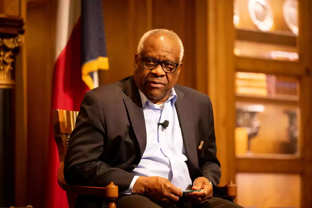 Older man with glasses and a dark jacket sitting on a wooden chair in a formal setting with a flag and bookshelves in the background Older man with glasses and a dark jacket sitting on a wooden chair in a formal setting with a flag and bookshelves in the background