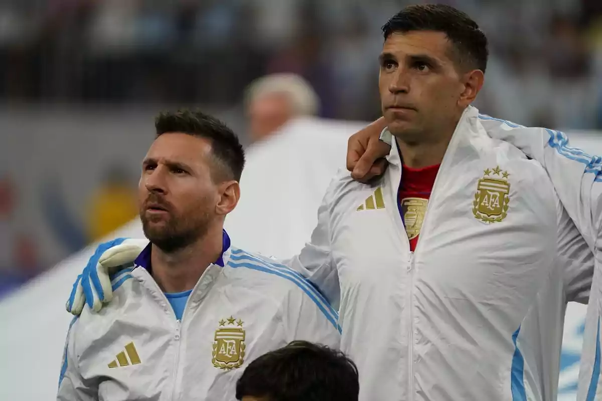 Dos jugadores de fútbol con chaquetas blancas de la selección argentina, uno con guantes, están de pie juntos en un estadio.