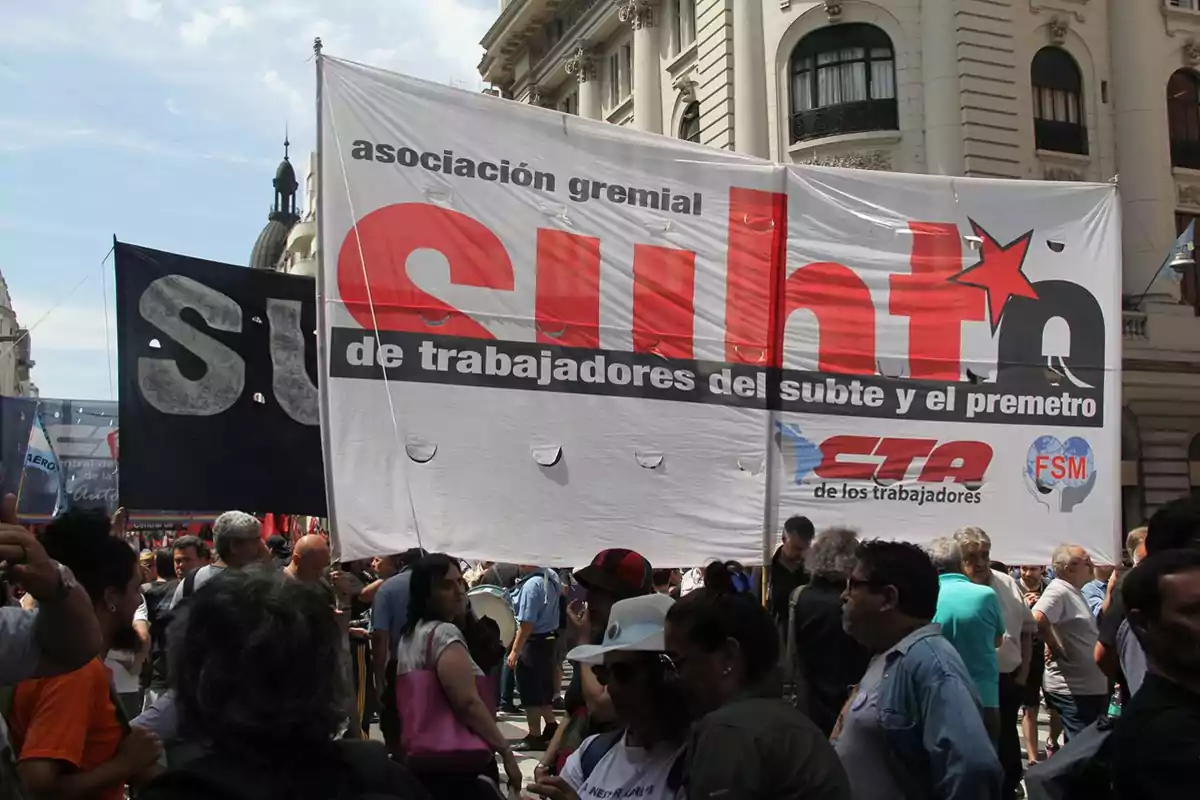 Un grupo de personas participa en una manifestación en la calle, sosteniendo una gran pancarta de la asociación gremial de trabajadores del subte y el premetro.