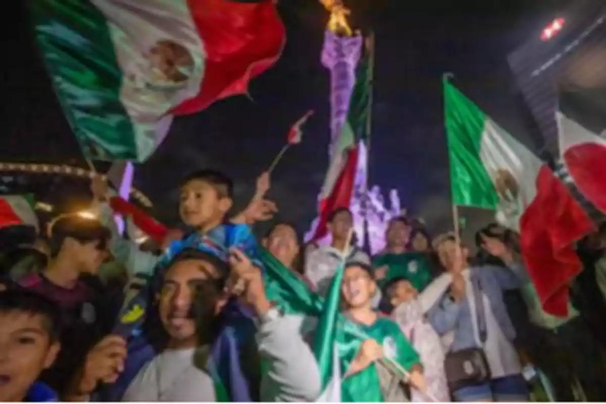 Un grupo de personas celebra con banderas mexicanas frente a un monumento iluminado por la noche.