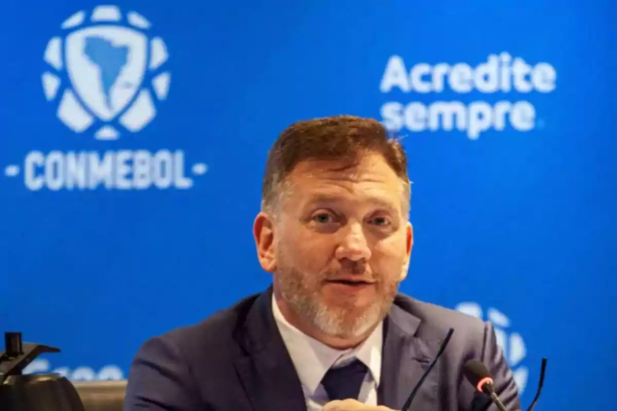 Hombre de traje hablando frente a un micrófono con fondo azul de CONMEBOL y el texto Acredite sempre Hombre de traje hablando frente a un micrófono con fondo azul de CONMEBOL y el texto Acredite sempre