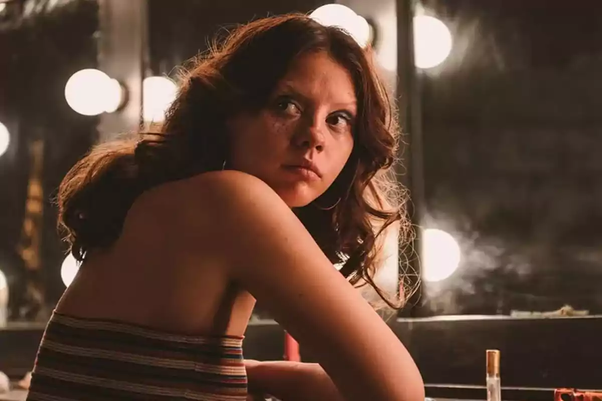 Joven con cabello ondulado y expresión seria sentada frente a un espejo iluminado con luces redondas Joven con cabello ondulado y expresión seria sentada frente a un espejo iluminado con luces redondas