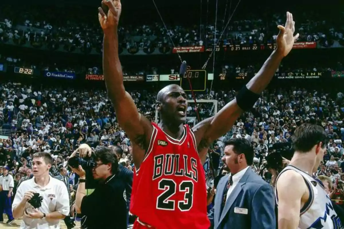 Un jugador de baloncesto con el uniforme de los Bulls levanta los brazos en señal de victoria mientras la multitud lo rodea en un estadio lleno. Un jugador de baloncesto con el uniforme de los Bulls levanta los brazos en señal de victoria mientras la multitud lo rodea en un estadio lleno.