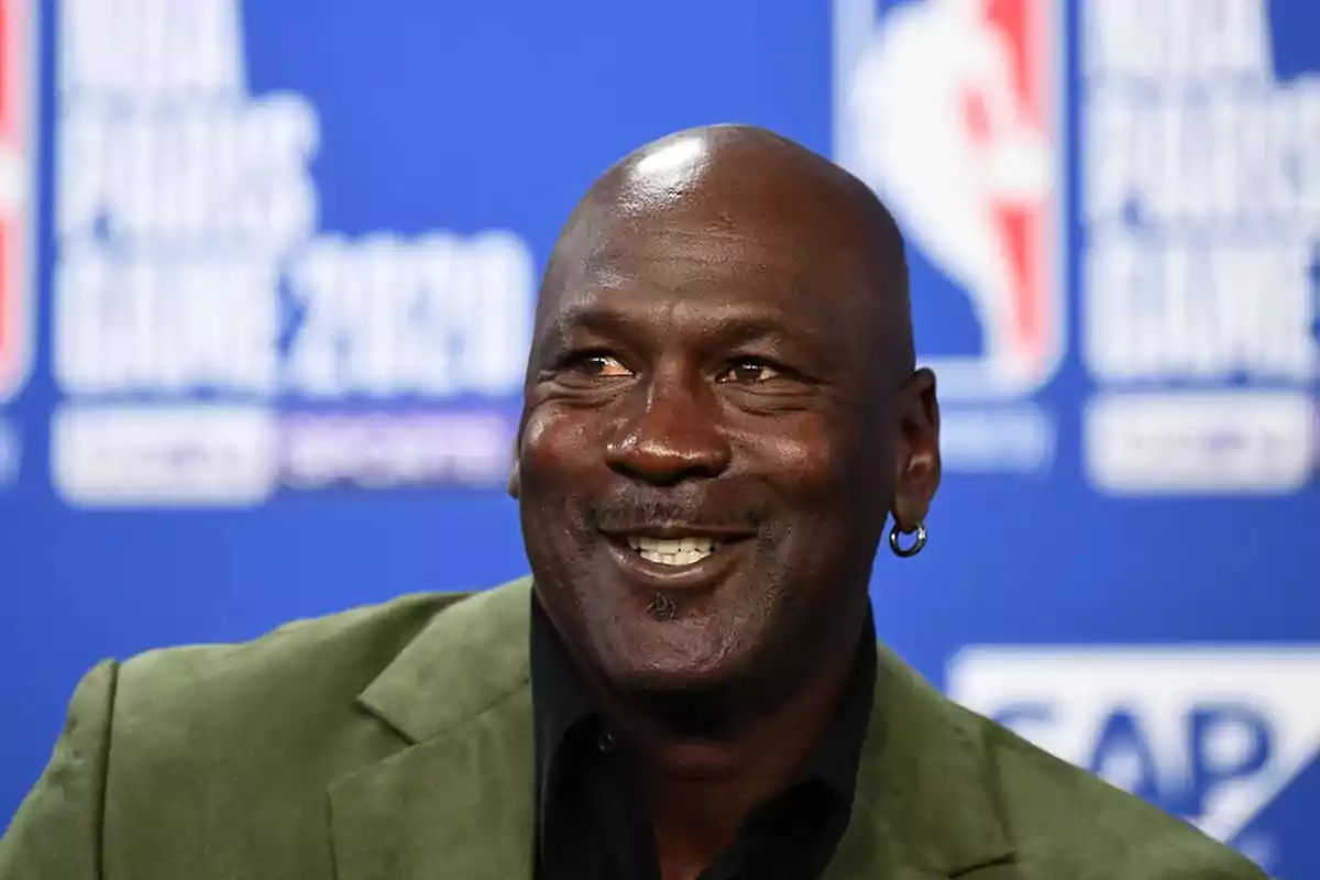 Un hombre sonriente con un pendiente en la oreja y un fondo azul con logotipos de la NBA.