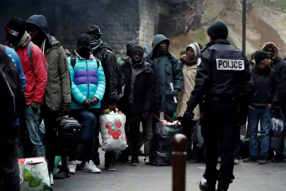 Un grupo de personas con abrigos y bolsas se encuentra en fila mientras un oficial de policía observa.