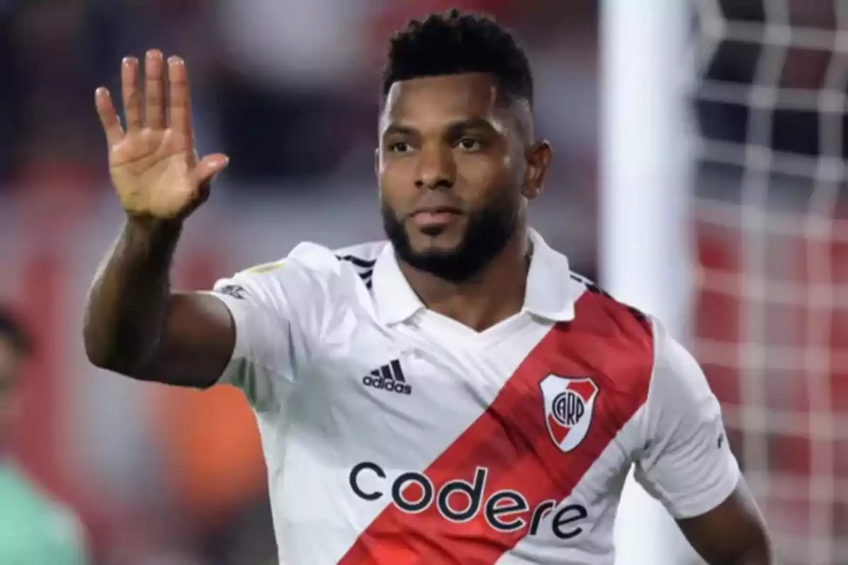 Un jugador de fútbol con uniforme blanco y rojo levanta la mano en señal de saludo en un estadio.