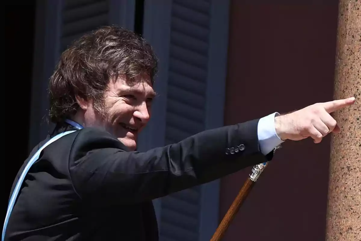 A man in a dark suit smiles while pointing forward with his finger, holding a cane in the other hand.