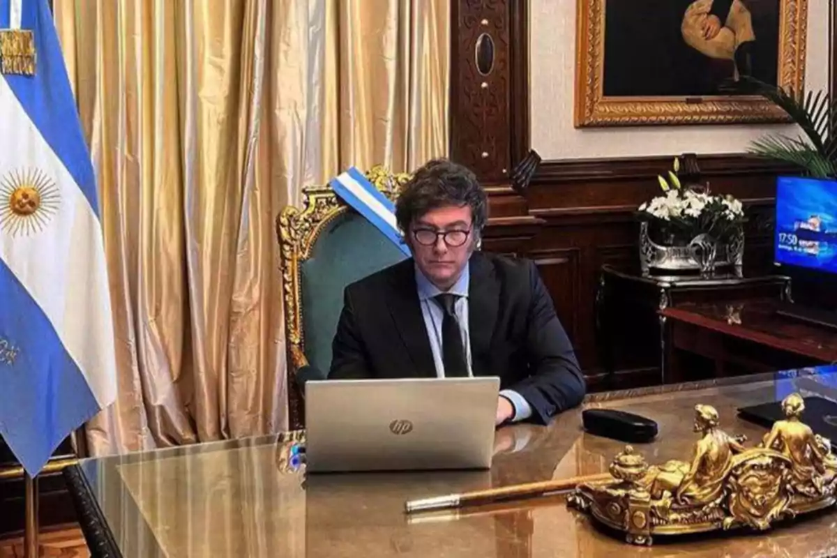 Un hombre con gafas y traje oscuro trabaja en una computadora portátil en un escritorio elegante con una bandera argentina y una decoración dorada.