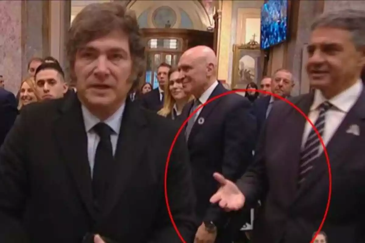 A brown-haired man in a dark suit is in the foreground while several people in formal attire are in the background in an elegant room, one of whom is surrounded by a red circle.