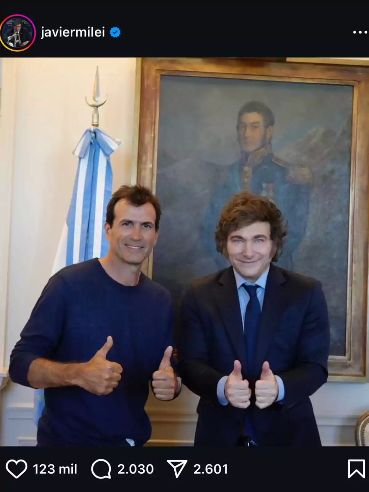 Dos hombres posan sonriendo con pulgares hacia arriba frente a un retrato y una bandera argentina.