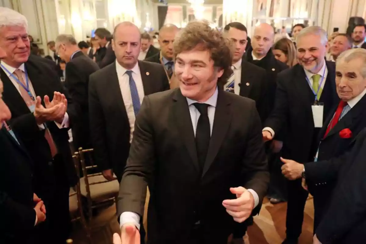 Un grupo de personas vestidas de traje en un evento social, con un hombre en el centro sonriendo y saludando.