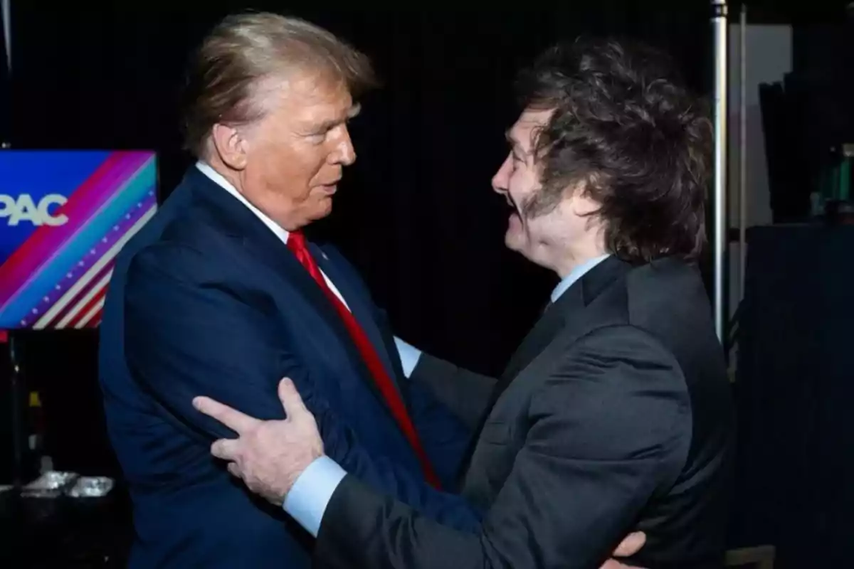 Dos personas se saludan cordialmente en un evento, una de ellas lleva un traje azul con corbata roja y la otra un traje oscuro.