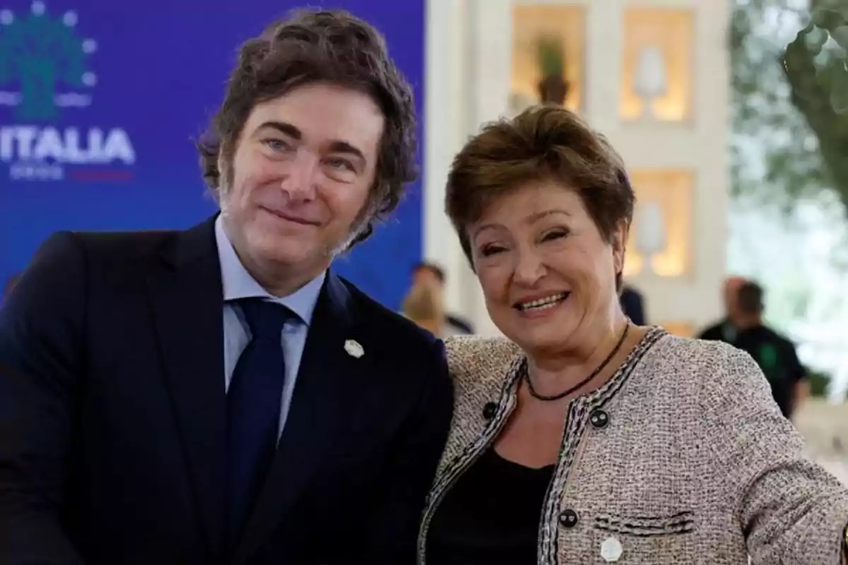 Dos personas sonrientes posan juntas en un evento formal con un fondo azul y decoración interior.